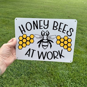 May include: White metal sign with black text that reads "Honey Bees At Work". The sign features a black and white bee illustration with yellow honeycomb graphics.