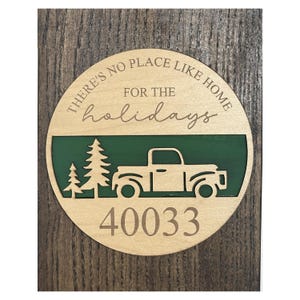 May include: A circular wooden sign featuring the engraved text THERE'S NO PLACE LIKE HOME FOR THE holidays. Below the text is a green horizontal panel with a cutout of a pickup truck and two pine trees. The number 40033 is engraved at the bottom.