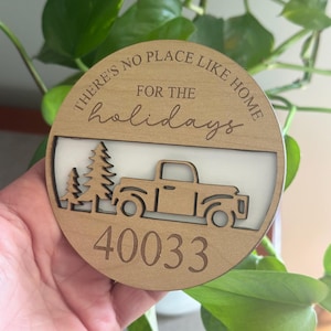 May include: A circular wooden ornament featuring the engraved text "THERE'S NO PLACE LIKE HOME FOR THE holidays" above a cutout design of two pine trees and a pickup truck, with the number "40033" engraved at the bottom.