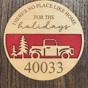 May include: A circular wooden ornament featuring the engraved text THERE'S NO PLACE LIKE HOME FOR THE holidays. Below the text is a red horizontal band with a cutout of a truck and two pine trees. The number 40033 is engraved at the bottom.