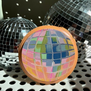May include: A round, painted wooden disc with a mirrored, mosaic-like design. The disc is painted in shades of pink, green, blue, and yellow. The disc is surrounded by two silver disco balls.