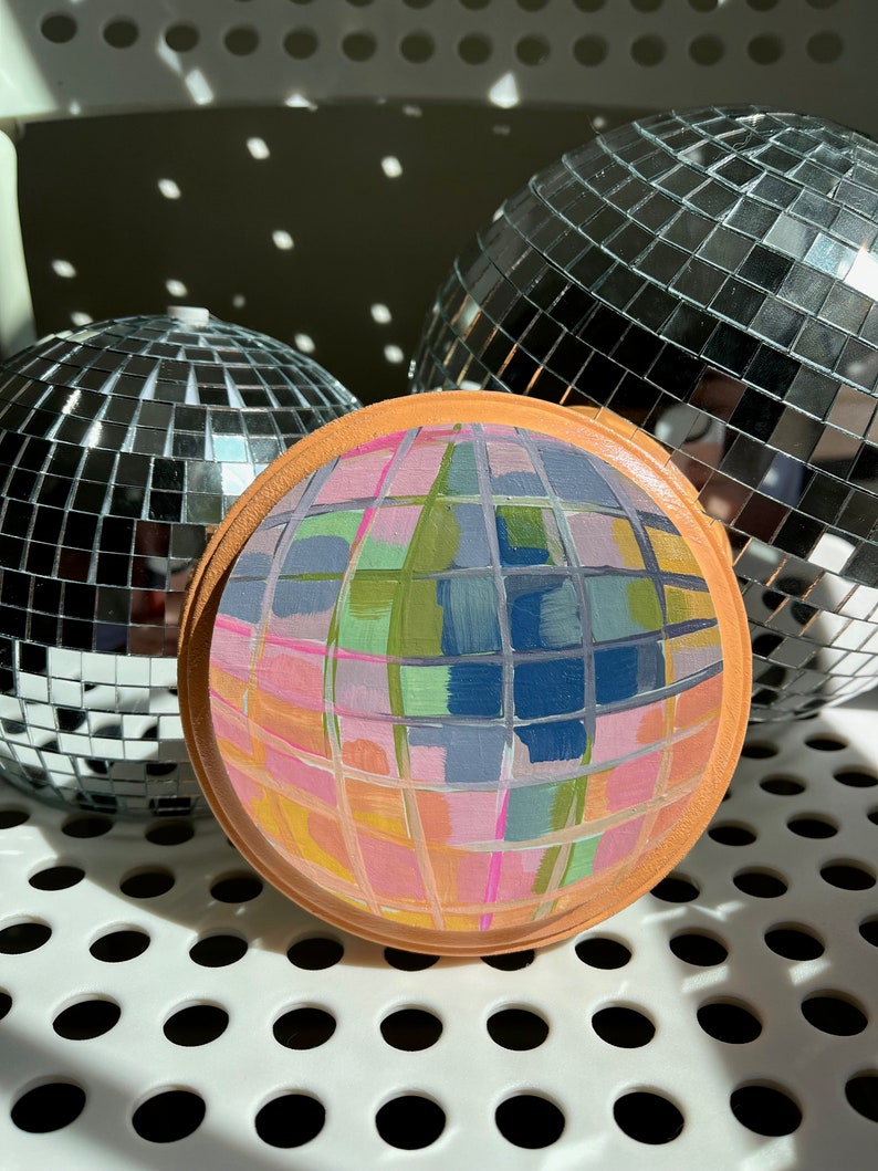 May include: A round, painted wooden disc with a mirrored, mosaic-like design. The disc is painted in shades of pink, green, blue, and yellow. The disc is surrounded by two silver disco balls.