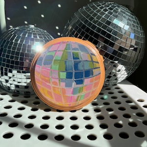 May include: Three disco balls, one with a painted design of colourful squares and rectangles, on a white surface with a grid pattern.