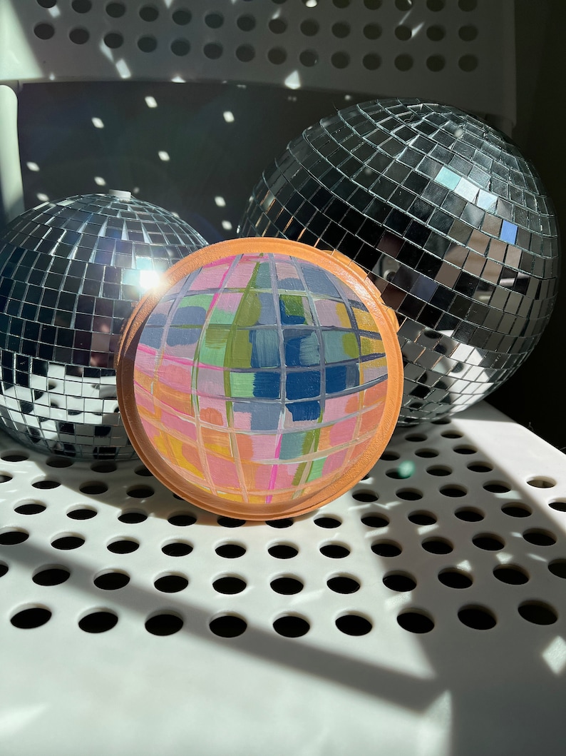 May include: Three disco balls, one with a painted design of colourful squares and rectangles, on a white surface with a grid pattern.