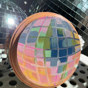 May include: A hand-painted disco ball with a geometric pattern in shades of pink, blue, green, and yellow. The ball is made of wood and has a brown rim.
