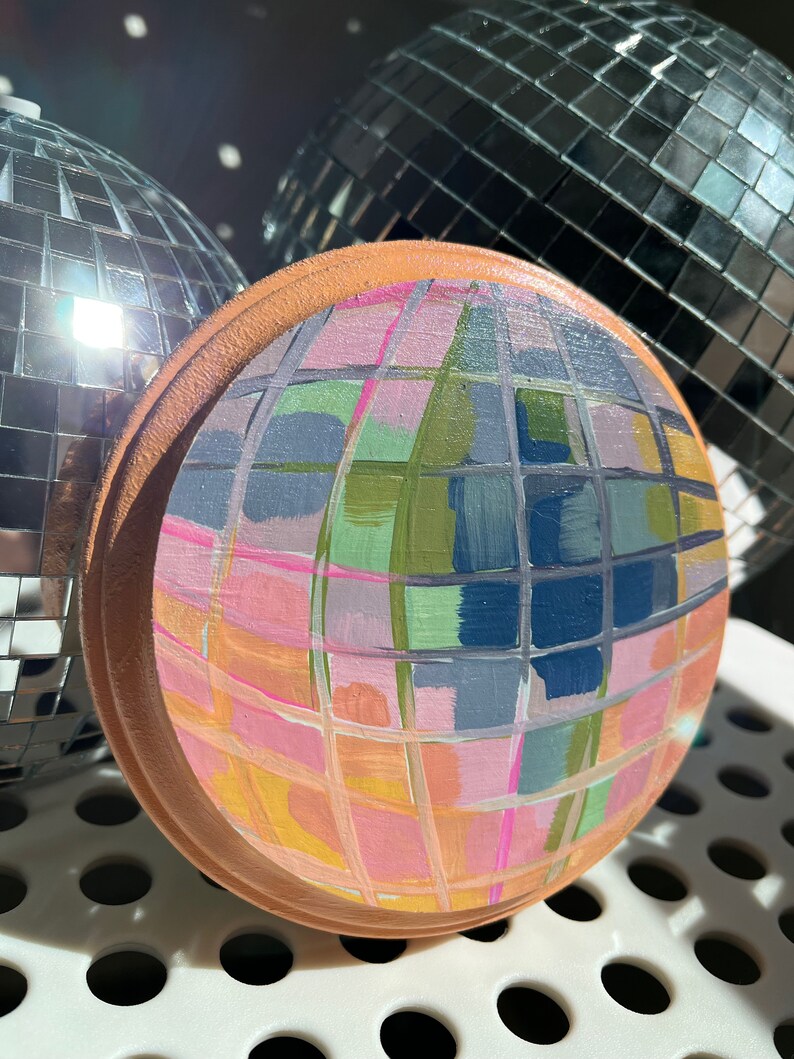 May include: A hand-painted disco ball with a geometric pattern in shades of pink, blue, green, and yellow. The ball is made of wood and has a brown rim.