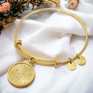 May include: Gold-coloured bangle bracelet with a round charm featuring a star map design. The bracelet has multiple strands and small charms that read "HAND MADE" and a flag. The bracelet is displayed on a white, textured surface.