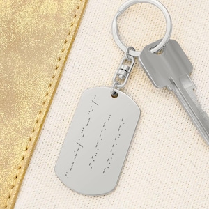 Peut inclure: Un porte-clés argenté en forme de plaque d'identification avec du texte en braille. Le porte-clés est attaché à un porte-clés argenté et à une clé. Le fond est un tissu beige clair et un matériau doré semblable à du cuir.