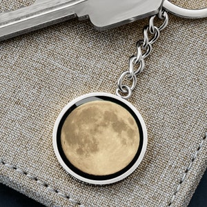 May include: A silver keychain with a round charm featuring a detailed image of the moon in shades of gray and yellow.