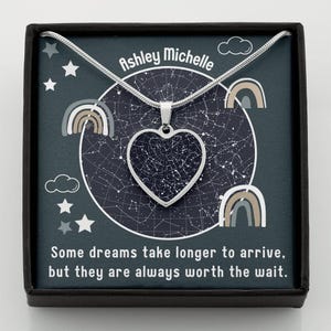 May include: A silver-toned heart-shaped pendant necklace featuring a star map design, presented in a dark blue box with the text Ashley Michelle and the quote Some dreams take longer to arrive, but they are always worth the wait, with rainbow icons.