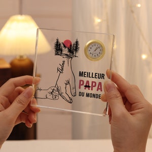 May include: A clear acrylic plaque with a black and white illustration of a hand holding a smaller hand. The text "Thelma" is written on the larger hand. The text "MEILLEUR PAPA DU MONDE" is written below the hands. A small gold clock is attached to the plaque.