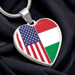 May include: Heart-shaped pendant with a silver-tone metal frame. The pendant features the American flag on one side and the Hungarian flag on the other. The pendant hangs from a silver-tone chain.
