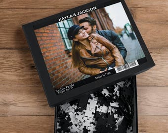 Peut inclure: Une boîte noire avec une photo d'un couple sur le devant. La photo montre une femme avec une veste en cuir marron et un homme avec une veste noire. La boîte indique "KAYLA & JACKSON" et "Fabriqué aux États-Unis". La boîte indique également "8 x 10 in Puzzle 120 pcs Ages 9+"