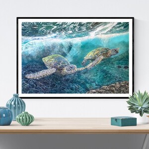 May include: Two green sea turtles swim through turquoise water with white foamy waves above them. The turtles are swimming in a coral reef with a dark blue background.