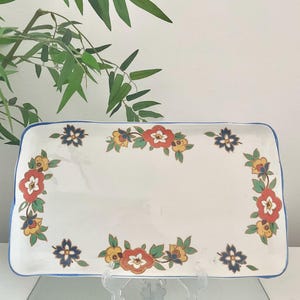 May include: A rectangular ceramic serving tray with a white base and a blue rim. The tray features a repeating floral pattern along the edges, consisting of red, yellow, and dark blue flowers with green leaves arranged in a symmetrical border design.