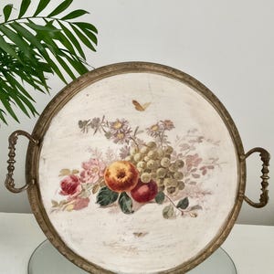 May include: Round serving tray with a painted floral and fruit design. The tray has a gold-coloured metal rim and handles. The artwork includes pink roses, grapes, and apples. The tray is approximately 35.5 cm in diameter.