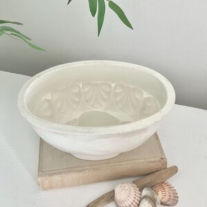 May include: A white ceramic bundt cake mould with a scalloped interior design. The mould is resting on a beige hardcover book, accompanied by a piece of driftwood and several small, light-coloured seashells arranged in a cluster nearby.