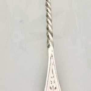 May include: A silver-toned metal fork featuring a three-pronged head, a slender twisted handle, and a wide, ornate base with intricate cut-out scrollwork and leaf-like engravings. The metal surface is polished and reflective throughout.