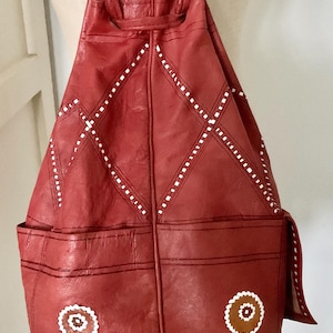 May include: Red leather backpack with white stitched diamond and dot patterns. Features two front pockets and decorative circular patches. Drawstring closure and adjustable straps.