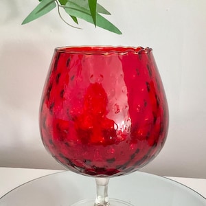 May include: A large, round, red glass goblet-style vase with a textured, dimpled surface. The vessel features a wide, bulbous bowl that tapers into a clear glass stem and a circular, transparent glass base, creating a distinct silhouette.