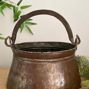 May include: A round, hammered copper pot featuring a wide, open top with a scalloped rim and a sturdy, arched metal handle attached to two side loops. The exterior surface displays a weathered, dark brown patina with visible metallic texture.