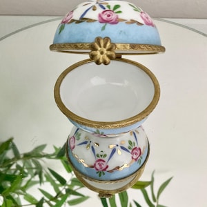 May include: A small, porcelain trinket box with a blue and white floral design. The box is in the shape of an egg and has a hinged lid. The lid is open, revealing the empty interior of the box.