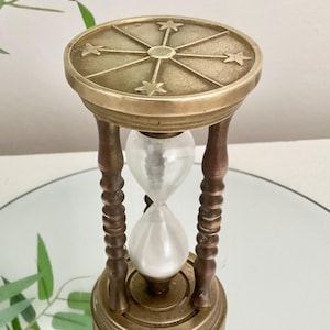 May include: An hourglass featuring a brass frame with a circular top plate engraved with four stars and radiating lines. The structure includes four turned wooden pillars connecting the top and base, enclosing a clear glass bulb filled with white sand.