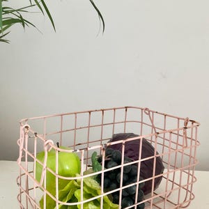 May include: A pink wire basket containing green lettuce, a green apple, a bunch of dark grapes, and a purple cabbage. The basket has a rectangular shape with a grid design and is on a white surface.