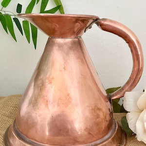 May include: A vintage copper jug with a wide, flared spout and a rounded handle. The jug has a smooth, polished surface and a slightly tarnished patina.