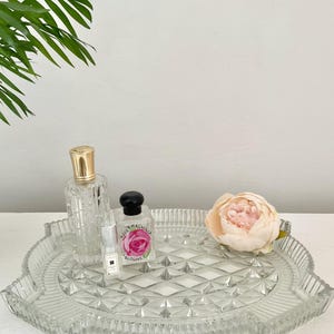 May include: A clear glass tray with a diamond pattern holds perfume bottles and a pink flower. One perfume bottle has a gold cap, another has a black cap and a pink rose label. A small perfume bottle is also on the tray.