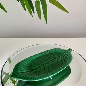 May include: A green ceramic leaf-shaped dish with a raised edge and a detailed vein pattern. The dish is sitting on a mirrored surface.