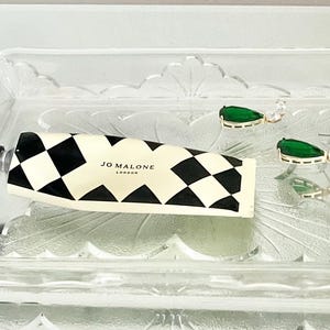 May include: A clear glass tray contains a tube of Jo Malone London hand cream with a black and white chequered pattern. Two gold-toned earrings with green teardrop-shaped stones and clear accents are also in the tray.