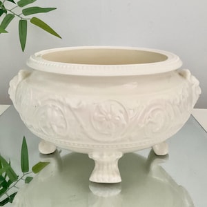 May include: A cream-coloured ceramic bowl featuring an embossed floral and scrollwork pattern around the exterior. The bowl has a wide circular opening, two side handles, and stands on three short, decorative feet with a beaded rim detail.