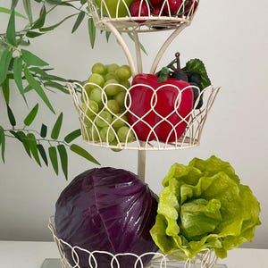 May include: A white metal three-tiered stand with a decorative design. The top tier holds a green apple and red cherries. The middle tier holds green grapes and a red apple. The bottom tier holds a red cabbage and green lettuce.