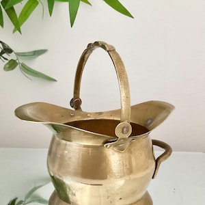 May include: A polished brass bucket with a wide, flared rim and a rounded body. It features a tall, arched handle attached with circular rivets and a smaller, fixed side handle. The metal surface shows a warm, golden metallic finish throughout.