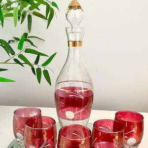 May include: A set of six ruby red glass decanter and shot glasses with gold accents and etched floral designs. The glasses are arranged on a mirrored tray.