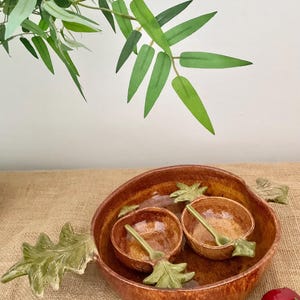 May include: A large, round, brown glazed ceramic serving bowl with two green leaf-shaped handles, containing two smaller matching bowls, each with a small green ceramic spoon resting inside, all featuring a textured, glossy finish.