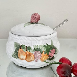 May include: White ceramic sugar bowl with a lid and a small spoon. The bowl is decorated with a fruit design featuring pears, grapes, and strawberries. The lid is topped with a strawberry-shaped knob.