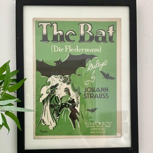 May include: A framed vintage poster for the operetta "The Bat" (Die Fledermaus) by Johann Strauss. The poster features a green background with black bats and a couple in period dress. The text "Walizes" is handwritten in black.