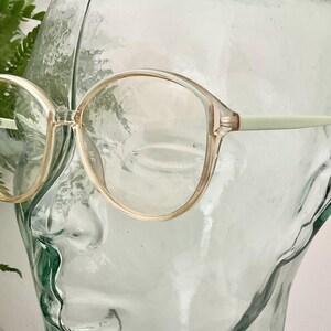 May include: Vintage spectacles with a light beige frame and clear lenses. The round frames have a subtle cat-eye shape. The arms are a pale green colour. The glasses are displayed on a clear glass head.