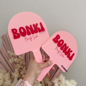 Puede incluir: Dos abanicos de mano rosas con letras rojas que dicen "BONKI Beauty Salon". Los abanicos se sostienen contra un fondo de flores secas y una pared gris claro. Los abanicos tienen un mango.