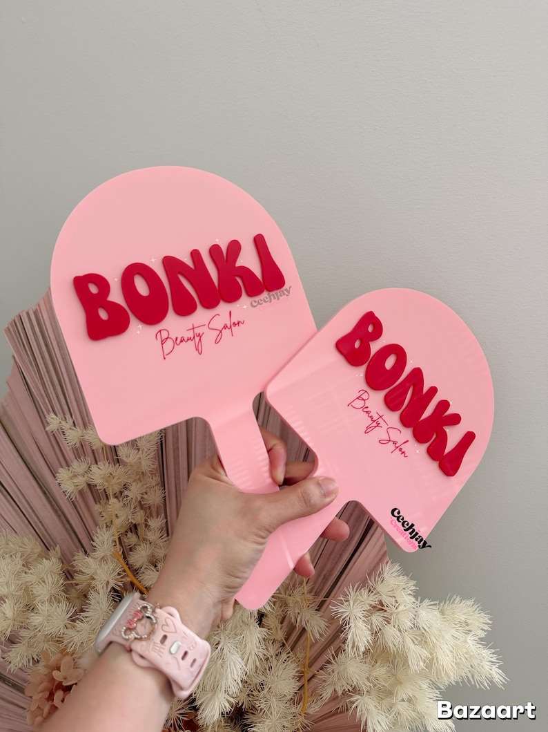 Puede incluir: Dos abanicos de mano rosas con letras rojas que dicen "BONKI Beauty Salon". Los abanicos se sostienen contra un fondo de flores secas y una pared gris claro. Los abanicos tienen un mango.
