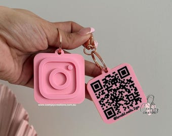 Puede incluir: Dos llaveros rosas de acrílico. Uno tiene forma de logotipo de Instagram, el otro tiene un código QR. Ambos tienen anillos dorados. El llavero de Instagram tiene la dirección del sitio web www.ceehjaycreations.com.au.