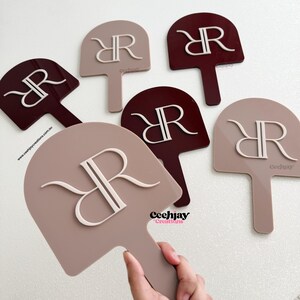 Puede incluir: Un conjunto de abanicos decorativos en tonos topo y burdeos, cada uno con un monograma "RR" estilizado en blanco. Los abanicos tienen forma de paleta con un asa. La marca "Ceechjay Creations" es visible en algunos abanicos.