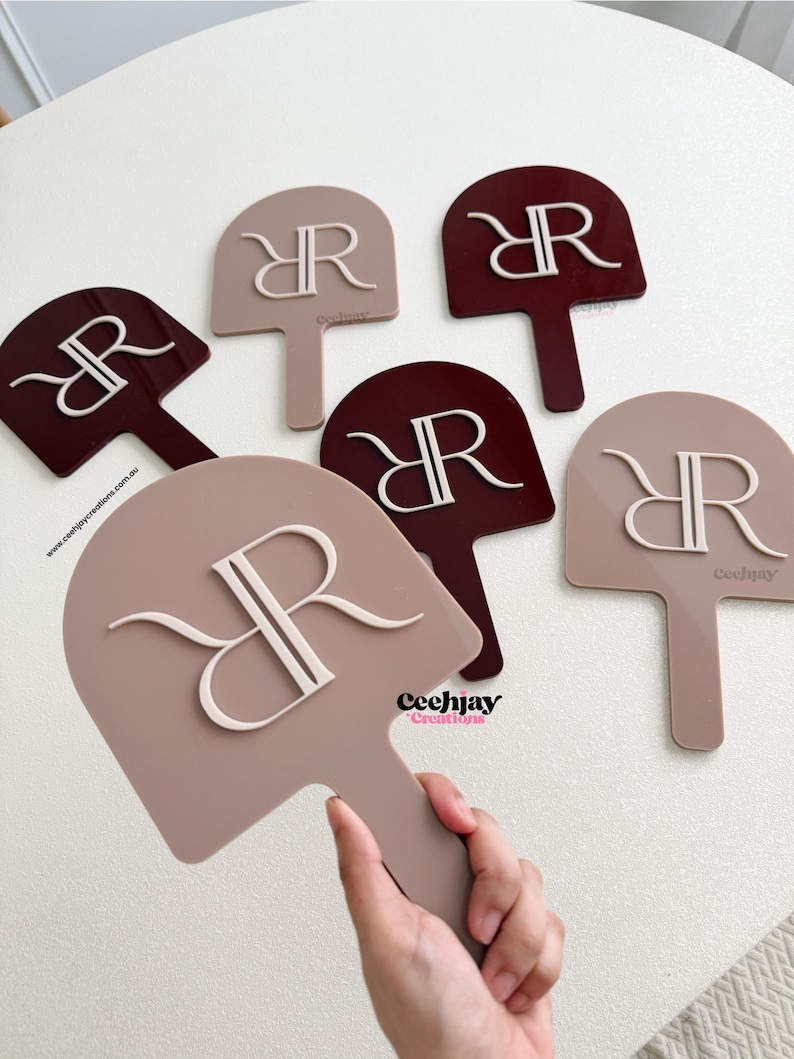 Puede incluir: Un conjunto de abanicos decorativos en tonos topo y burdeos, cada uno con un monograma "RR" estilizado en blanco. Los abanicos tienen forma de paleta con un asa. La marca "Ceechjay Creations" es visible en algunos abanicos.
