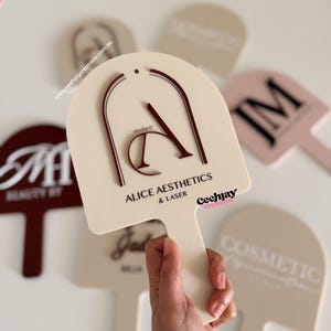 Puede incluir: Paleta de color crema con el texto "ALICE AESTHETICS & LASER" en negro y un dise&ntilde;o "A" burdeos. La paleta se sostiene en una mano, con otras paletas similares en el fondo. La marca "Ceehjay Creations" tambi&eacute;n es visible.