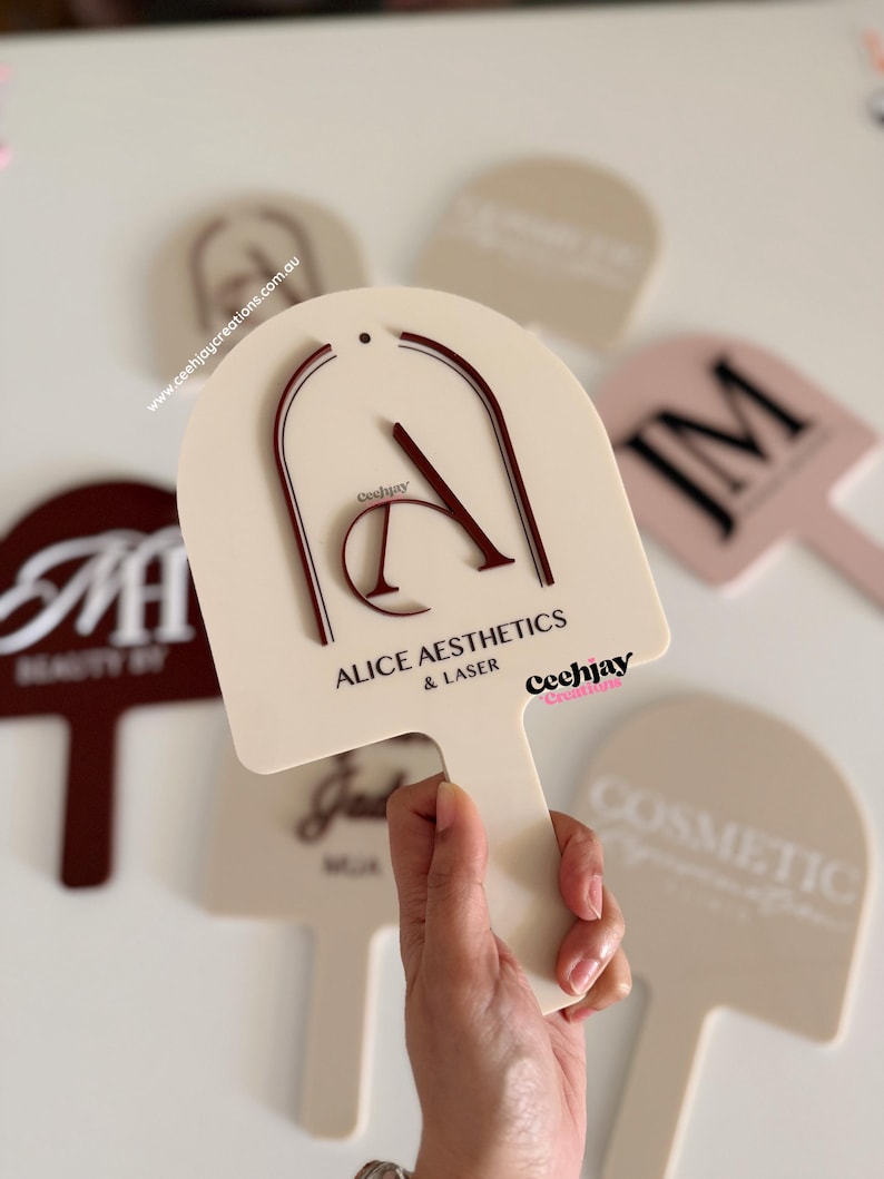 Puede incluir: Paleta de color crema con el texto "ALICE AESTHETICS & LASER" en negro y un dise&ntilde;o "A" burdeos. La paleta se sostiene en una mano, con otras paletas similares en el fondo. La marca "Ceehjay Creations" tambi&eacute;n es visible.