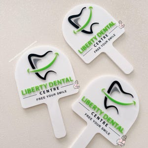 Puede incluir: Tres letreros blancos en forma de paleta con el texto "LIBERTY DENTAL CENTRE - FREE YOUR SMILE" en verde. Cada letrero presenta un dise&ntilde;o de diente estilizado con acentos negros, grises y verdes. Los letreros est&aacute;n dispuestos sobre una superficie clara.