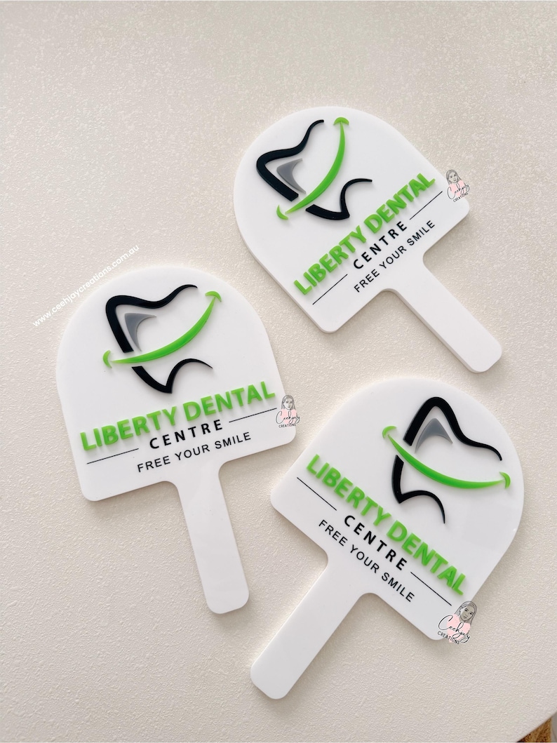 Puede incluir: Tres letreros blancos en forma de paleta con el texto "LIBERTY DENTAL CENTRE - FREE YOUR SMILE" en verde. Cada letrero presenta un dise&ntilde;o de diente estilizado con acentos negros, grises y verdes. Los letreros est&aacute;n dispuestos sobre una superficie clara.