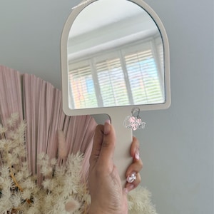 Puede incluir: Un espejo de mano color crema con la parte superior arqueada se sostiene frente a una pared gris claro. El mango del espejo est&aacute; integrado en su marco. La direcci&oacute;n del sitio web "www.ceejaycreations.com.au" est&aacute; impresa en la parte superior del espejo.